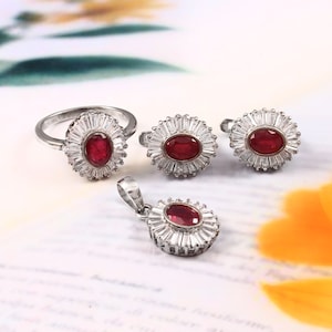 May include: A silver ring, a pair of earrings, and a pendant necklace, all featuring a red oval gemstone surrounded by a halo of smaller white gemstones.