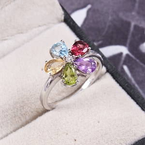 Op de afbeelding: Een zilveren ring met een bloemmotief met zes gekleurde edelstenen: lichtblauw, geel, groen, paars en rood. De ring ligt in een wit fluwelen doosje.