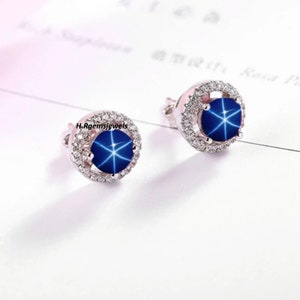 Peut inclure: Une paire de boucles d'oreilles en argent avec une pierre précieuse saphir étoilé bleue entourée d'un halo de petites pierres blanches.