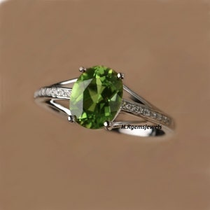 Può includere: Un anello d'argento con una grande pietra di peridoto ovale. Il peridoto è di un verde brillante ed è il punto focale dell'anello. La fascia si divide in due, con un lato ornato da piccole pietre preziose trasparenti. Il testo "H. Rgemsjewels" è visibile.