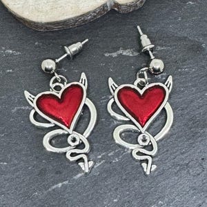 May include: Silver-coloured earrings with a red heart design, devil horns, and a tail. The heart is outlined in silver and filled with red. The earrings have a post-back closure.