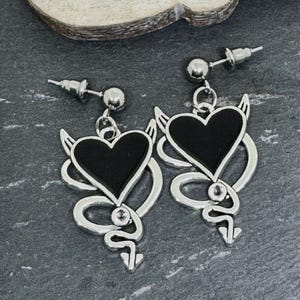 May include: A pair of silver earrings with a black heart design, devil horns, and a stylized tail. The earrings have a post-back closure and a small ball accent. The earrings are displayed on a dark grey surface.