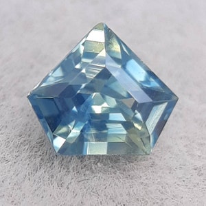 Peut inclure: Une pierre précieuse bleu clair taillée en forme de pentagone. La gemme présente de multiples facettes qui réfléchissent la lumière, créant un effet scintillant. La pierre est posée sur un fond blanc texturé.