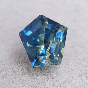 May include: A faceted gemstone with a pentagonal shape. The gem displays a range of blue and green hues, with a brilliant, reflective surface. The gemstone is set against a white background.