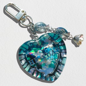 May include: A heart-shaped keychain with a blue and green resin design. The resin contains glitter, iridescent flakes, and a silver dolphin charm. The keychain also features a silver chain with three silver fish charms and a silver flower charm. The keychain says "H.S.UAN" in silver lettering.