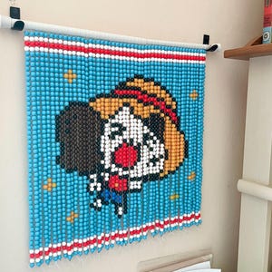 May include: A blue beaded curtain featuring a cartoon dog wearing a red hat and a white shirt. The dog has a red nose and black eyes. The curtain is hanging on a white rod.