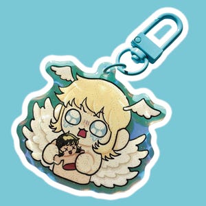 Op de afbeelding: Een glitterende sleutelhanger met een cartoon engel met blond haar en witte vleugels, die een kleinere figuur vasthoudt. De engel heeft een bedroefde uitdrukking met tranen. De sleutelhanger heeft een blauwe sluiting en een lichtblauwe achtergrond.