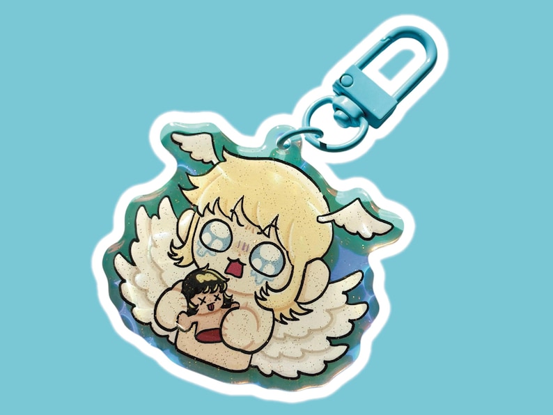 Op de afbeelding: Een glitterende sleutelhanger met een cartoon engel met blond haar en witte vleugels, die een kleinere figuur vasthoudt. De engel heeft een bedroefde uitdrukking met tranen. De sleutelhanger heeft een blauwe sluiting en een lichtblauwe achtergrond.