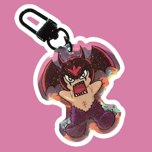 Op de afbeelding: Een sleutelhanger in cartoonstijl met een duivels karakter met rode hoorns en vleugels en een woeste uitdrukking. De sleutelhanger heeft een zwarte sluiting en een glinsterend, holografisch effect. Het karakter is paars en bruin met een kruissymbool.