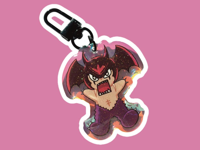Op de afbeelding: Een sleutelhanger in cartoonstijl met een duivels karakter met rode hoorns en vleugels en een woeste uitdrukking. De sleutelhanger heeft een zwarte sluiting en een glinsterend, holografisch effect. Het karakter is paars en bruin met een kruissymbool.