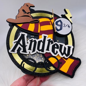 Può includere: Un topper per torta rotondo nero e oro con una sciarpa rossa e gialla, un cappello da mago marrone, un paio di occhiali rotondi e un fulmine con il numero 9 3/4. Il nome Andrew è scritto in lettere bianche sul topper.
