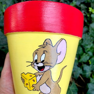 May include: Hand-painted flower pot featuring Jerry from Tom and Jerry cartoon. The pot has a yellow base with Jerry holding cheese, a red rim, and a brown and white design. The pot is designed for planting flowers.
