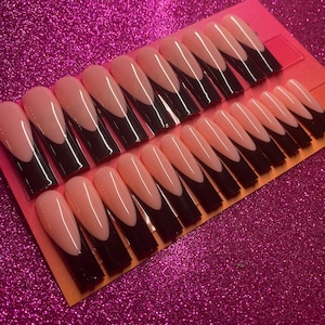 Könnte beinhalten: Ein Set mit 24 falschen Nägeln mit einer Nude-Basis und schwarzen Spitzen. Die Nägel sind in einer Reihe auf einem rosa und orangefarbenen Hintergrund angeordnet.