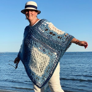 Op de afbeelding: Een vrouw draagt een blauw-witte gebreide poncho met een witte gebreide hoed op een strand.