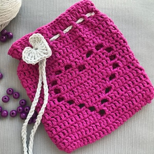 May include: A crocheted pink drawstring bag with a white heart-shaped embellishment. The bag has a white crocheted drawstring.