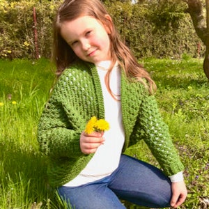 May include: A green crocheted cardigan with a lacy pattern. The cardigan is being worn by a young person in a grassy field.