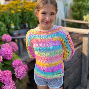Puede incluir: Un suéter de ganchillo a rayas de arcoíris con mangas largas. El suéter presenta una mezcla de colores pastel, incluyendo rosa, amarillo, azul y verde. El suéter se usa con pantalones cortos grises.