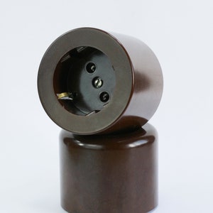 Peut inclure: Deux caches-prises électriques en bakélite marron empilés l'un sur l'autre. Le cache supérieur comporte trois trous pour les fils électriques.