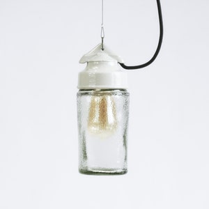 Könnte beinhalten: Eine weiße Keramik-Pendelleuchte mit einem klaren Glasschirm. Der Glasschirm hat eine strukturierte Oberfläche und eine Glühbirne ist im Inneren sichtbar.