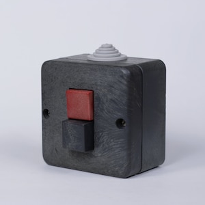 May include: A black plastic electrical switch with a red and black button. The switch has a gray plastic cover on top.