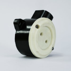 May include: A black electrical socket with a white faceplate. The socket has three holes and is secured with screws. The design is a vintage style, with a cylindrical body and a rectangular top.