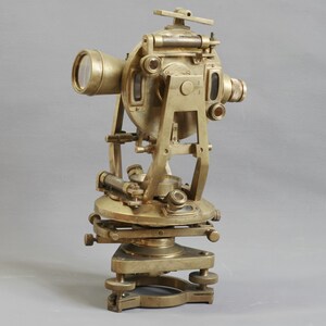 Puede incluir: Un teodolito de latón antiguo, un instrumento de topografía, con un telescopio, tornillos de nivelación y una base circular. El instrumento tiene un diseño complejo con múltiples lentes y perillas de ajuste. El latón tiene una pátina cálida y envejecida.