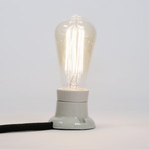 Może przedstawiać: Przezroczysta żarówka szklana z białym ceramicznym gniazdkiem i czarnym kablem. Żarówka ma włókno z wieloma drutami.