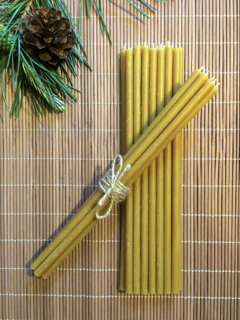 May include: A bundle of 12 natural beeswax candles tied together with twine. The candles are a light yellow color and are arranged on a woven bamboo mat.