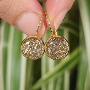 Può includere: Un paio di orecchini pendenti rotondi con una montatura in metallo dorato e un inserto in glitter dorato scintillante e testurizzato. Gli orecchini sono sospesi a sottili ganci in filo metallico dorato curvo fissati sulla parte superiore.