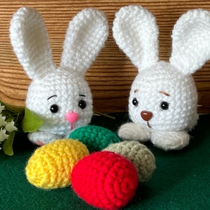 Puede incluir: Dos conejos de crochet blanco con ojos negros y narices marrones se sientan sobre una superficie verde con cuatro huevos de Pascua de crochet en amarillo, rojo, verde y marr&oacute;n.