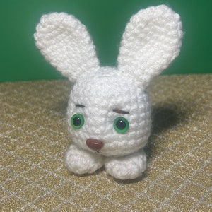 Puede incluir: Un conejito de crochet blanco con ojos verdes y una nariz marr&oacute;n. El conejito est&aacute; sentado sobre una superficie con un patr&oacute;n dorado y blanco.