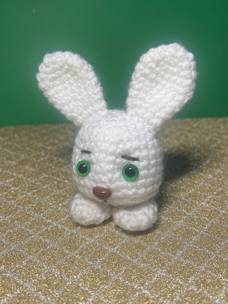 Puede incluir: Un conejito de crochet blanco con ojos verdes y una nariz marr&oacute;n. El conejito est&aacute; sentado sobre una superficie con un patr&oacute;n dorado y blanco.