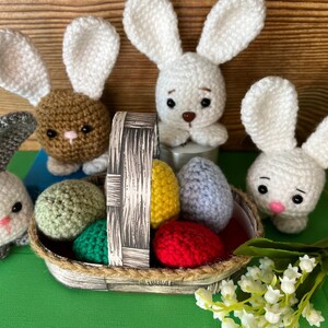 Puede incluir: Una cesta de crochet llena de huevos de Pascua coloridos, rodeada de cuatro conejos de crochet. Los conejos son blancos, marrones y grises. La cesta est&aacute; hecha de mimbre marr&oacute;n y tiene un asa de cuerda blanca.