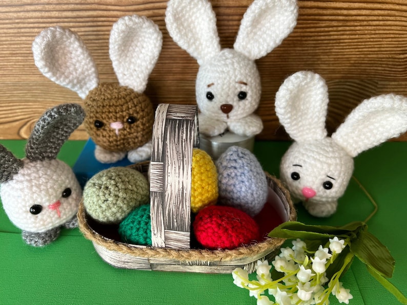 Puede incluir: Una cesta de crochet llena de huevos de Pascua coloridos, rodeada de cuatro conejos de crochet. Los conejos son blancos, marrones y grises. La cesta est&aacute; hecha de mimbre marr&oacute;n y tiene un asa de cuerda blanca.
