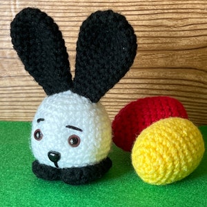 Puede incluir: Un conejito de crochet blanco con orejas negras y una nariz negra. El conejito est&aacute; sentado en una superficie verde con dos bolas de crochet, una roja y una amarilla.