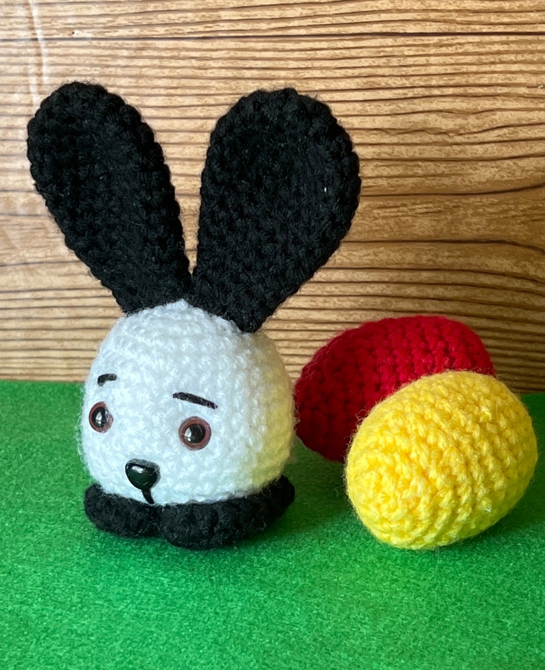 Puede incluir: Un conejito de crochet blanco con orejas negras y una nariz negra. El conejito est&aacute; sentado en una superficie verde con dos bolas de crochet, una roja y una amarilla.