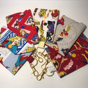 May include: A collection of fabric pouches and keychains featuring comic book superheroes. The pouches display images of Wonder Woman, Superman, and Batman. The keychains have gold-colored clasps and yellow fabric loops.