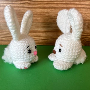 Puede incluir: Dos conejos de crochet blanco con ojos negros y narices rosas. Los conejos se miran y tienen una cola blanca y esponjosa.