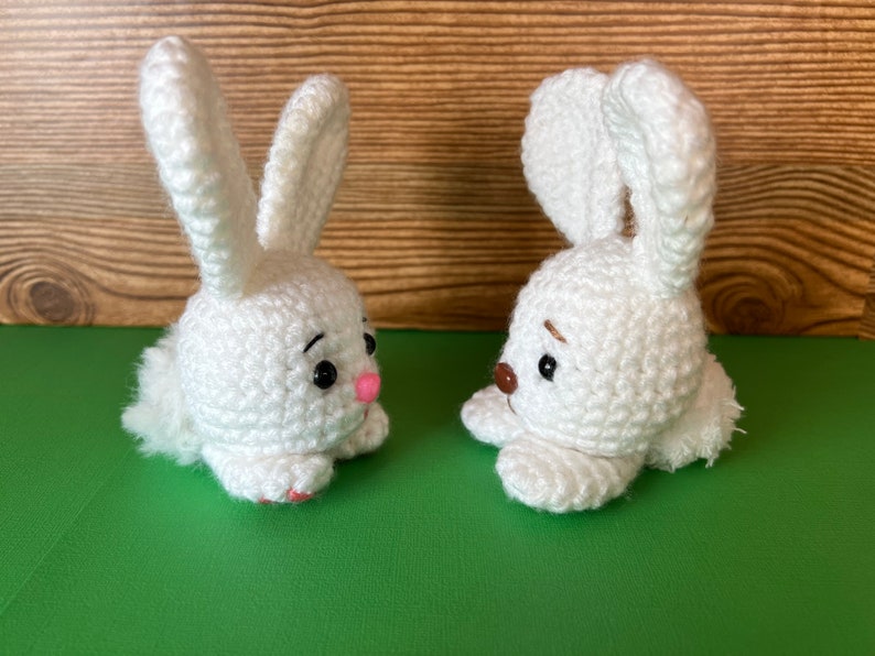 Puede incluir: Dos conejos de crochet blanco con ojos negros y narices rosas. Los conejos se miran y tienen una cola blanca y esponjosa.