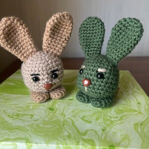 Puede incluir: Dos conejos de crochet, uno marr&oacute;n y otro verde, con caras bordadas y ojos negros. Los conejos est&aacute;n sentados sobre una superficie de m&aacute;rmol verde y blanco.