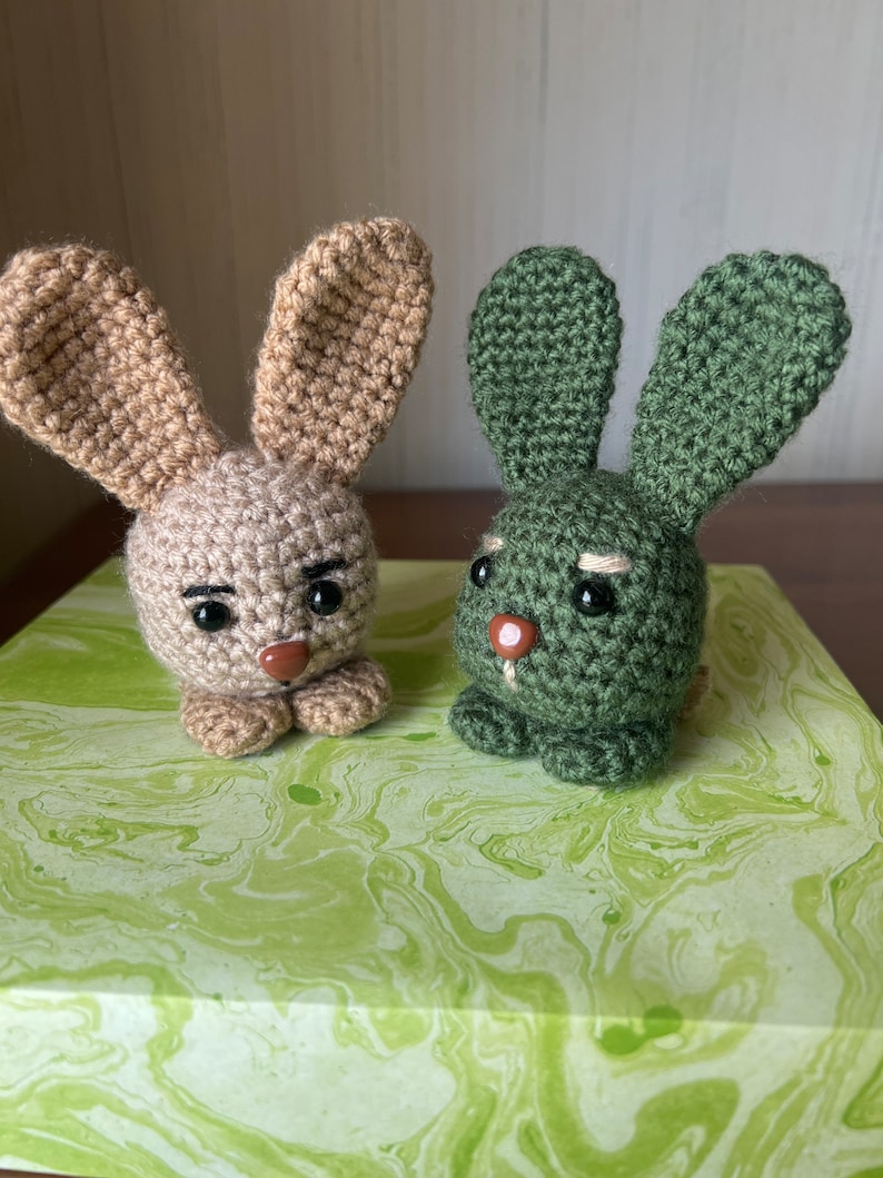 Puede incluir: Dos conejos de crochet, uno marr&oacute;n y otro verde, con caras bordadas y ojos negros. Los conejos est&aacute;n sentados sobre una superficie de m&aacute;rmol verde y blanco.