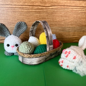 Puede incluir: Dos conejos de crochet, uno sosteniendo una cesta llena de huevos de Pascua de crochet de colores. La cesta est&aacute; hecha de papel tejido y tiene un patr&oacute;n marr&oacute;n y blanco.