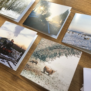 May include: A collection of greeting cards featuring winter landscape photographs. Scenes include a snow-covered forest, a road with a blue line, a steam train, Highland cattle in a snowy field, and sheep in a snowy field. The cards are displayed on a wooden surface.