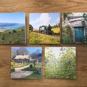 May include: Five square cards with scenic images. One shows a coastal view, another a steam train on tracks, a third a stone building with a teal door, a fourth a thatched-roof cottage, and the last, a field of purple and blue flowers.