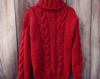 May include: A vibrant red turtleneck sweater with a cable knit design. The sweater is made of a thick, chunky knit material and hangs on a hanger against a wooden background. The sleeves and front panels feature a detailed cable pattern.