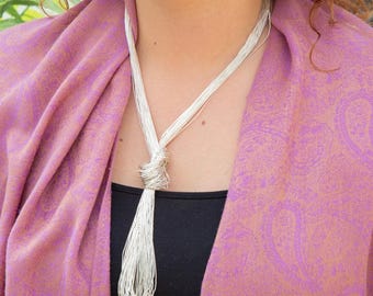 May include: A silver-toned necklace with multiple strands knotted at the center, cascading into a fringe. The necklace is worn over a black top and a pink and gold paisley patterned shawl.