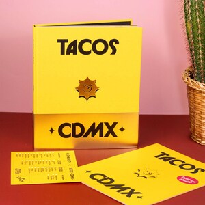 May include: A bright yellow hardcover book titled TACOS CDMX with a gold metallic bottom section and a sun icon. Beside it lies a yellow rectangular card and a yellow booklet, both featuring the same TACOS CDMX text and sun graphic.