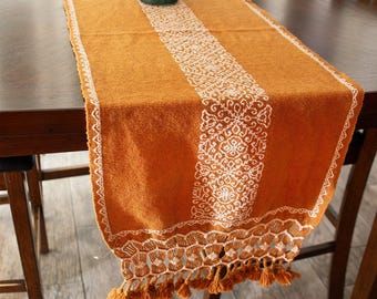 May include: A long, orange table runner with white embroidered floral designs and tassels. The runner is laid on a wooden table.