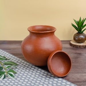May include: A terracotta-colored clay pot with a matching lid, set on a patterned cloth. The pot has a rounded body and a wide opening. A small plant in a brown pot sits in the background.