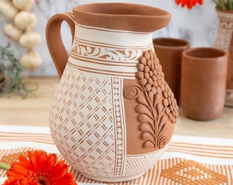 May include: A terracotta pitcher with a handle and a wide spout. The pitcher features a white and brown geometric design, with a raised floral relief on the side. Two matching brown cups are in the background.